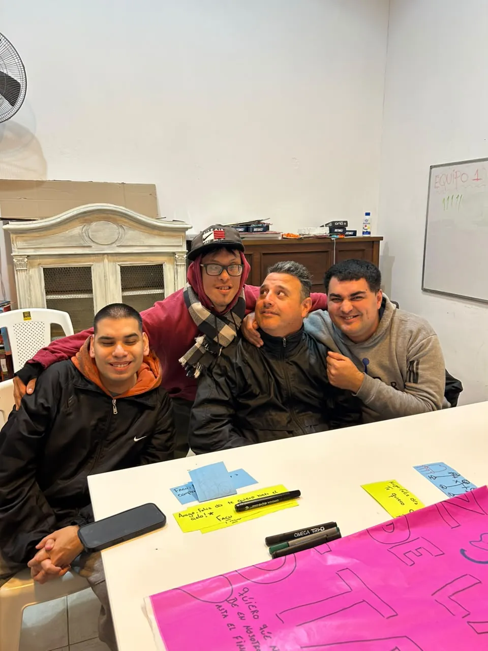 Cuatro concurrentes abrazados y sonriendo alrededor de una mesa de trabajo del centro